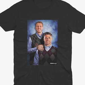 May include: A black t-shirt featuring a graphic of two men wearing sweaters. The men are smiling and standing next to each other. The background is a blue and white gradient.