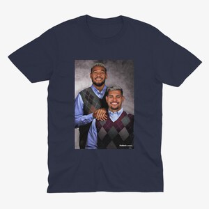 Puede incluir: Camiseta azul marino con un retrato de dos personas con chalecos de rombos sobre camisas azules. La imagen tiene el texto "Futbol Classics". La camiseta es de cuello redondo.