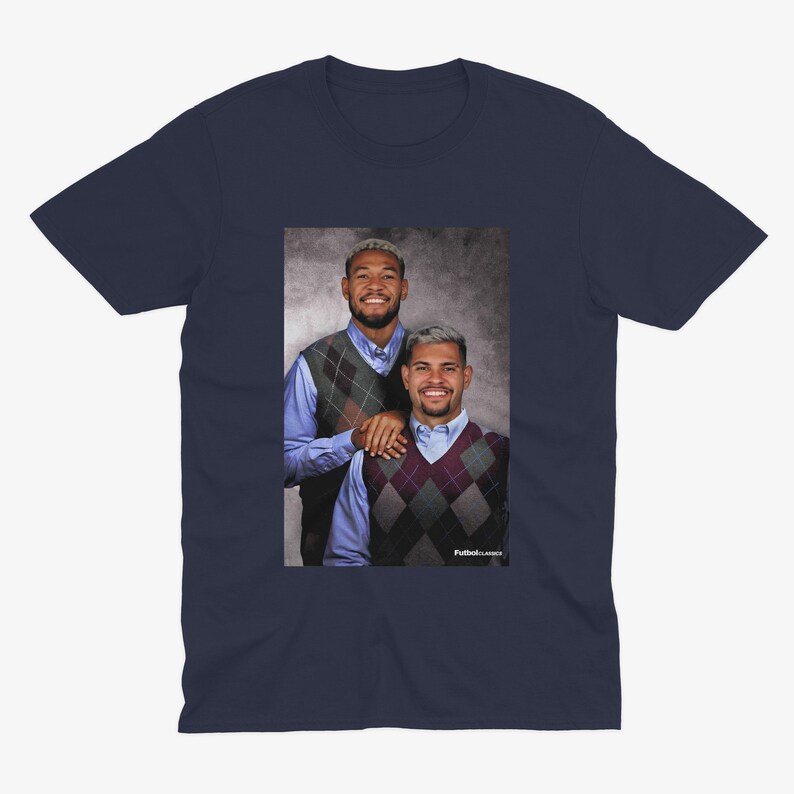 Puede incluir: Camiseta azul marino con un retrato de dos personas con chalecos de rombos sobre camisas azules. La imagen tiene el texto "Futbol Classics". La camiseta es de cuello redondo.