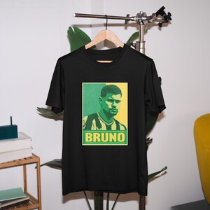 K&ouml;nnte beinhalten: Schwarzes T-Shirt mit einer Grafik eines Mannes mit gr&uuml;nen und gelben Akzenten. Das Wort "BRUNO" wird in einem gr&uuml;nen Rechteck darunter angezeigt. Das T-Shirt h&auml;ngt an einem Holzkleiderb&uuml;gel.
