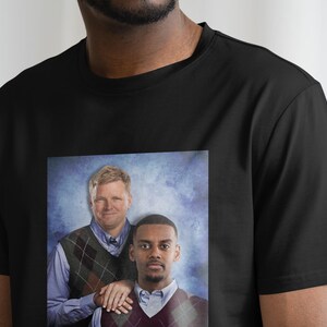May include: A black t-shirt with a printed image of two men wearing sweaters. The men are standing in front of a blue and white background. The man on the left is wearing a brown and white patterned sweater. The man on the right is wearing a red and white patterned sweater.