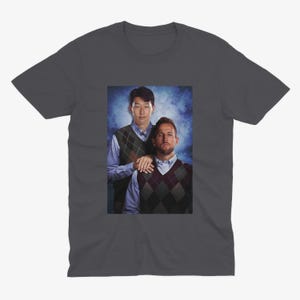 Op de afbeelding: Een donkergrijs T-shirt met een foto van twee mannen in truien. De man links draagt een blauw shirt met een groen-bruine geruite trui eroverheen. De man rechts draagt een blauw shirt met een zwart-witte geruite trui eroverheen. De mannen staan voor een blauw-witte achtergrond.