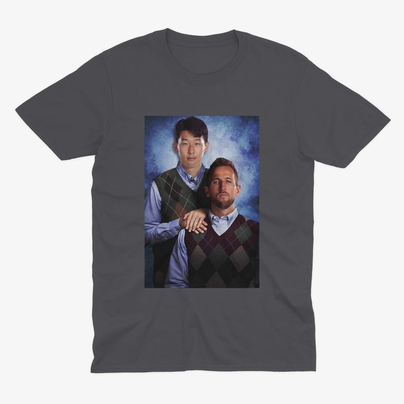 Op de afbeelding: Een donkergrijs T-shirt met een foto van twee mannen in truien. De man links draagt een blauw shirt met een groen-bruine geruite trui eroverheen. De man rechts draagt een blauw shirt met een zwart-witte geruite trui eroverheen. De mannen staan voor een blauw-witte achtergrond.