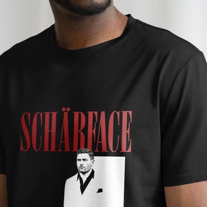 May include: Black t-shirt with the word "SCH&Auml;RFACE" in red, above a black and white image of a man in a suit. The t-shirt is made of a soft material.