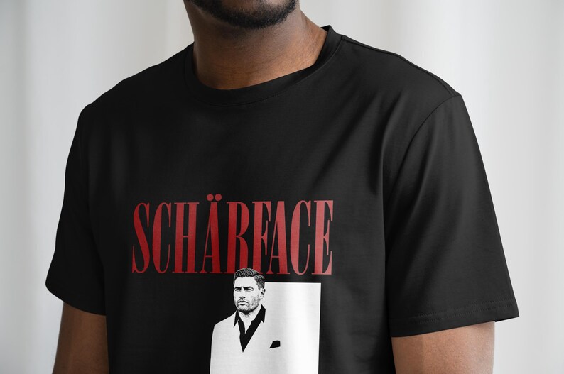 May include: Black t-shirt with the word "SCH&Auml;RFACE" in red, above a black and white image of a man in a suit. The t-shirt is made of a soft material.