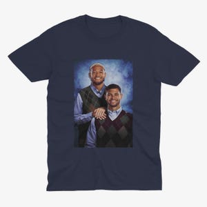 K&ouml;nnte beinhalten: Ein marineblaues T-Shirt mit einem Foto von zwei M&auml;nnern, die Pullover tragen. Die M&auml;nner stehen nebeneinander und l&auml;cheln. Der Hintergrund ist ein blauer und wei&szlig;er Farbverlauf.