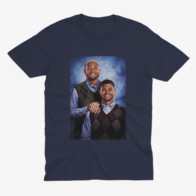K&ouml;nnte beinhalten: Ein marineblaues T-Shirt mit einem Foto von zwei M&auml;nnern, die Pullover tragen. Die M&auml;nner stehen nebeneinander und l&auml;cheln. Der Hintergrund ist ein blauer und wei&szlig;er Farbverlauf.