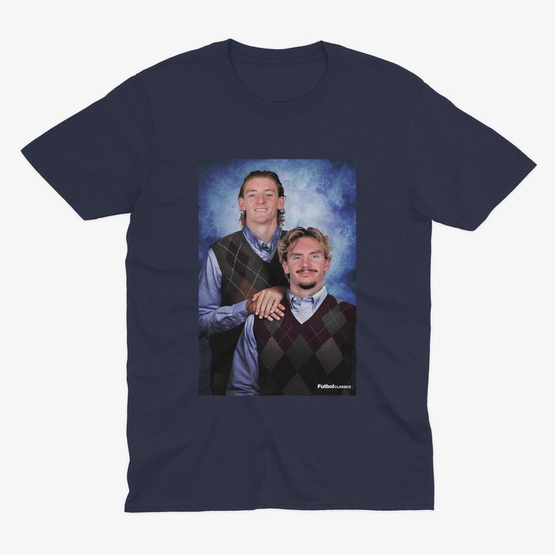 May include: Navy blue t-shirt featuring a portrait of two people wearing argyle vests over button-down shirts. The image has a vintage aesthetic with a blue background. The text "Futbol Classics" is visible in the bottom right corner.