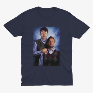 Op de afbeelding: Een marineblauw t-shirt met een foto van twee mannen die truien dragen. De man links draagt een groen-witte trui met een patroon en de man rechts draagt een bruin-witte trui met een patroon.