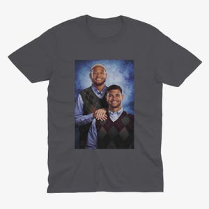 K&ouml;nnte beinhalten: Ein dunkelgraues T-Shirt mit einem Foto von zwei M&auml;nnern, die Pullover tragen. Die M&auml;nner stehen vor einem blauen und wei&szlig;en Hintergrund. Der Mann links tr&auml;gt ein blaues Hemd und einen braunen und wei&szlig;en gemusterten Pullover. Der Mann rechts tr&auml;gt ein wei&szlig;es Hemd und einen schwarzen und wei&szlig;en gemusterten Pullover.