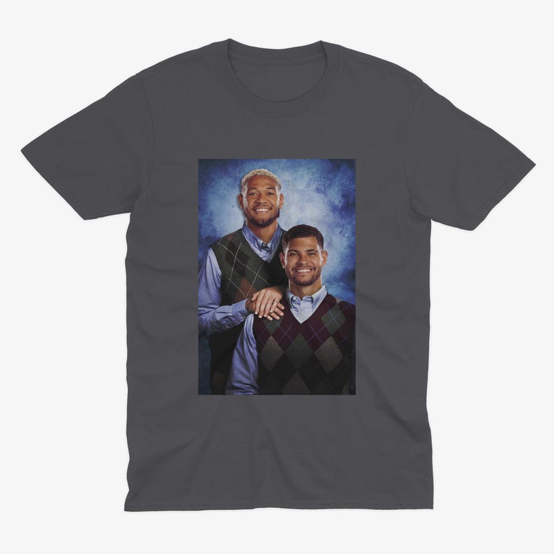 K&ouml;nnte beinhalten: Ein dunkelgraues T-Shirt mit einem Foto von zwei M&auml;nnern, die Pullover tragen. Die M&auml;nner stehen vor einem blauen und wei&szlig;en Hintergrund. Der Mann links tr&auml;gt ein blaues Hemd und einen braunen und wei&szlig;en gemusterten Pullover. Der Mann rechts tr&auml;gt ein wei&szlig;es Hemd und einen schwarzen und wei&szlig;en gemusterten Pullover.