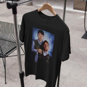 Op de afbeelding: Zwart T-shirt met een grafische print van twee mannen, een met een blauw-witte gepatroneerde trui en de ander met een blauw-witte gepatroneerde trui. De achtergrond is een blauw-witte gradi&euml;nt.
