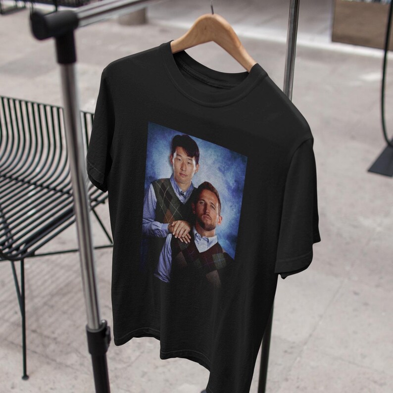 Op de afbeelding: Zwart T-shirt met een grafische print van twee mannen, een met een blauw-witte gepatroneerde trui en de ander met een blauw-witte gepatroneerde trui. De achtergrond is een blauw-witte gradi&euml;nt.