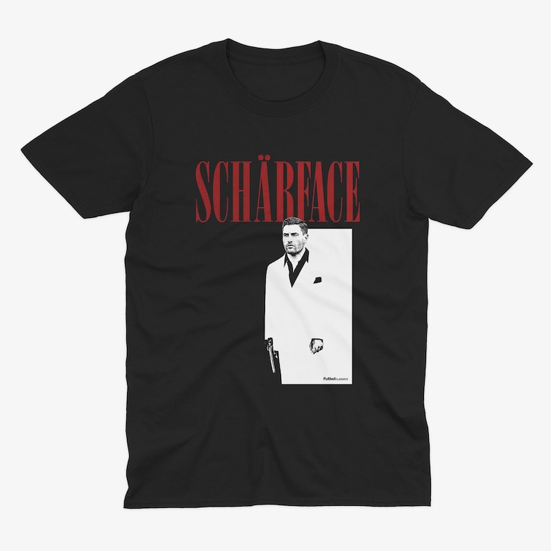 May include: Black t-shirt featuring the word "SCH&Auml;RFACE" in red, above a black and white image of a man in a white suit. The man is holding a gun. The shirt is a crew neck.