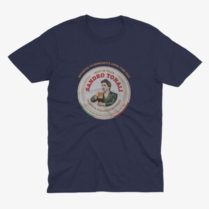 Pu&ograve; includere: Una maglietta blu navy con un'etichetta in stile vintage che presenta un'immagine di un calciatore dei cartoni animati che tiene una birra. L'etichetta recita "Sandro Tonali, Made in Italia, Centrocampista italiano di alta qualit&agrave;, Importato a Newcastle Upon Tyne 2023".