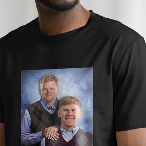 Pu&ograve; includere: Una maglietta nera con un'immagine stampata di due uomini con un maglione a motivi blu e bianchi. Gli uomini sorridono e stanno uno accanto all'altro.