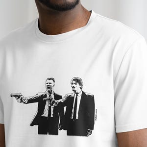 May include: White t-shirt featuring a black and white graphic of two men in suits pointing guns. The image is a stylized illustration, with one man yelling. The shirt is made of a soft material.
