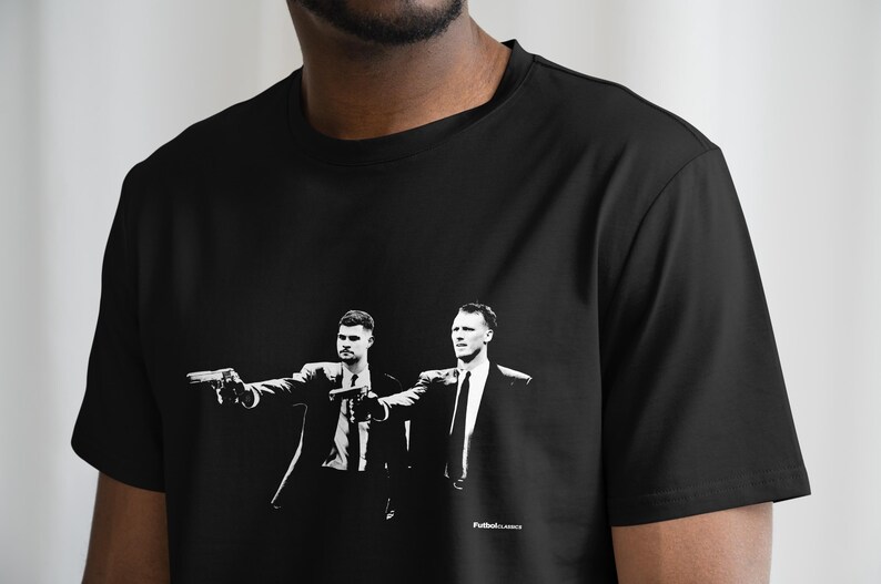 May include: Black t-shirt featuring a black and white graphic of two men in suits holding guns. The image is a stylized illustration. The text "Futbol.Lances" is printed at the bottom right of the graphic.