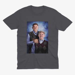 May include: A dark grey t-shirt featuring a photo of two people wearing jumpers. The photo is set against a blue and white background.