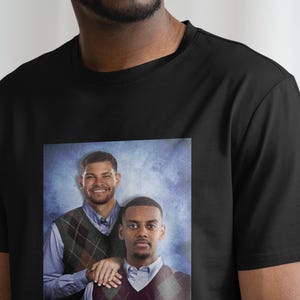 May include: Black t-shirt featuring a printed image of two men wearing sweaters and posing for a photograph. The background is a blue and white gradient.