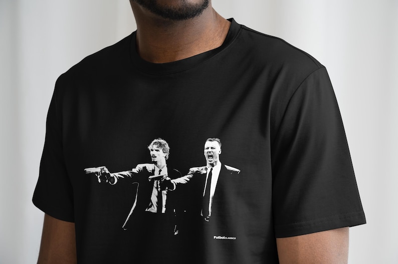 May include: Black t-shirt featuring a black and white graphic of two men in suits pointing guns. The image is a stylized illustration. The text "Futbol Classics" is printed at the bottom of the graphic.