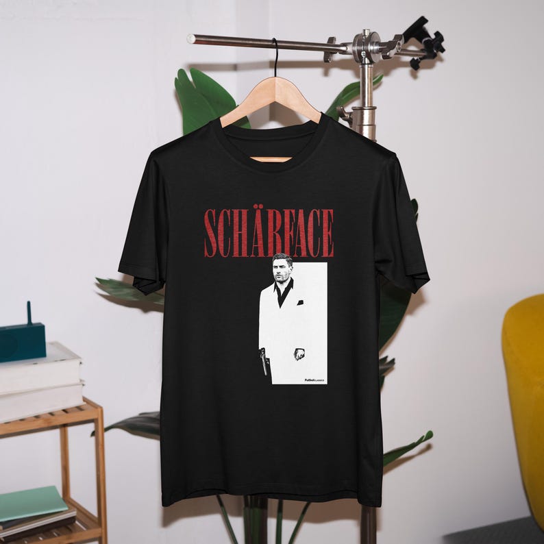 May include: Black t-shirt with the word "SCH&Auml;RFACE" in red, above a black and white graphic of a man in a white suit holding a gun. The shirt is hanging on a wooden hanger.