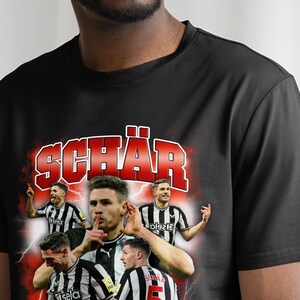 May include: Black t-shirt featuring a red and white graphic print of a football player with the name "SCH&Auml;R" in large red letters.
