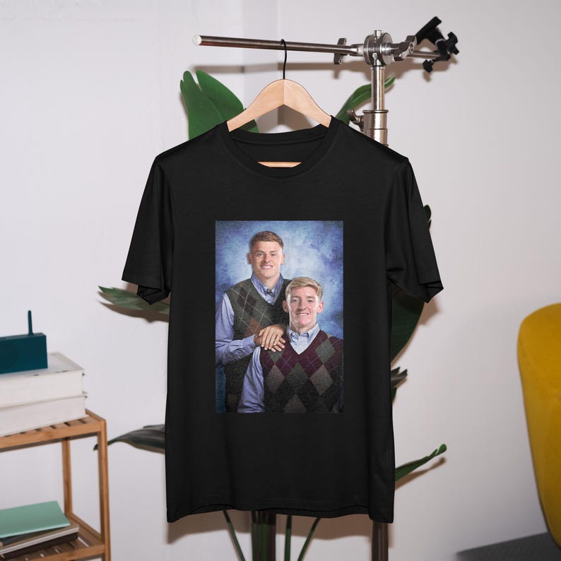 May include: Black t-shirt featuring a photograph of two men wearing sweaters. The men are standing side-by-side and smiling. The background is a blue and white gradient.