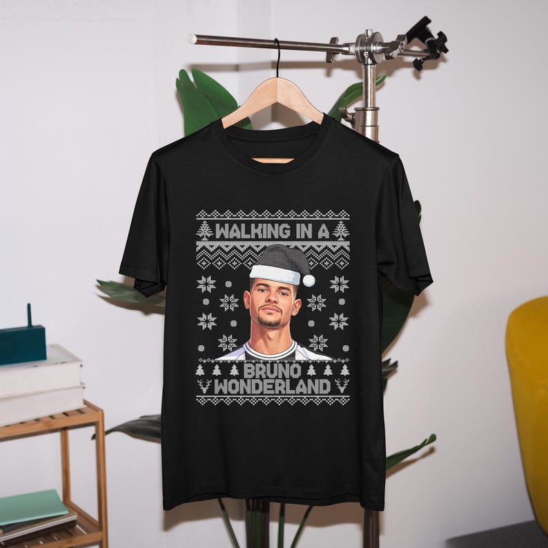 K&ouml;nnte beinhalten: Schwarzes T-Shirt mit festlichem Design. Es zeigt ein Portr&auml;t einer Person mit Weihnachtsm&uuml;tze, mit dem Text "WALKING IN A" &uuml;ber dem Portr&auml;t und "BRUNO WONDERLAND" darunter, zusammen mit Schneeflocken- und Weihnachtsbaumgrafiken.