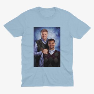 May include: Light blue t-shirt featuring a portrait of two men. The men are wearing argyle vests over button-down shirts. The background is a blurred blue and white.