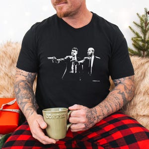 May include: Black V-neck t-shirt featuring a black and white graphic of two men in suits pointing guns. The shirt is worn by a person holding a green mug with the word "COFFEE" on it. The person is wearing red and black plaid pajama pants.