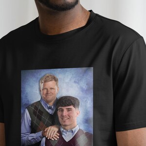 May include: A black t-shirt with a printed image of two men in sweaters. The men are standing in front of a blue and white background. The image is a portrait style photo.