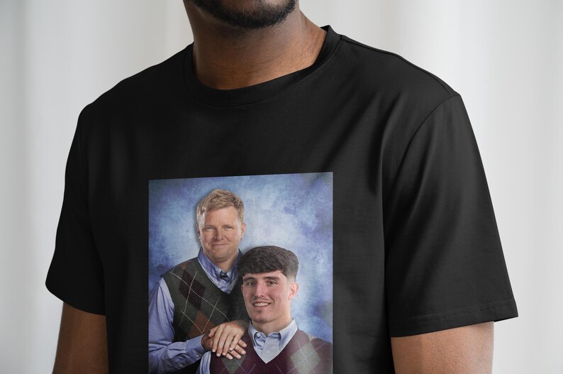 May include: A black t-shirt with a printed image of two men in sweaters. The men are standing in front of a blue and white background. The image is a portrait style photo.