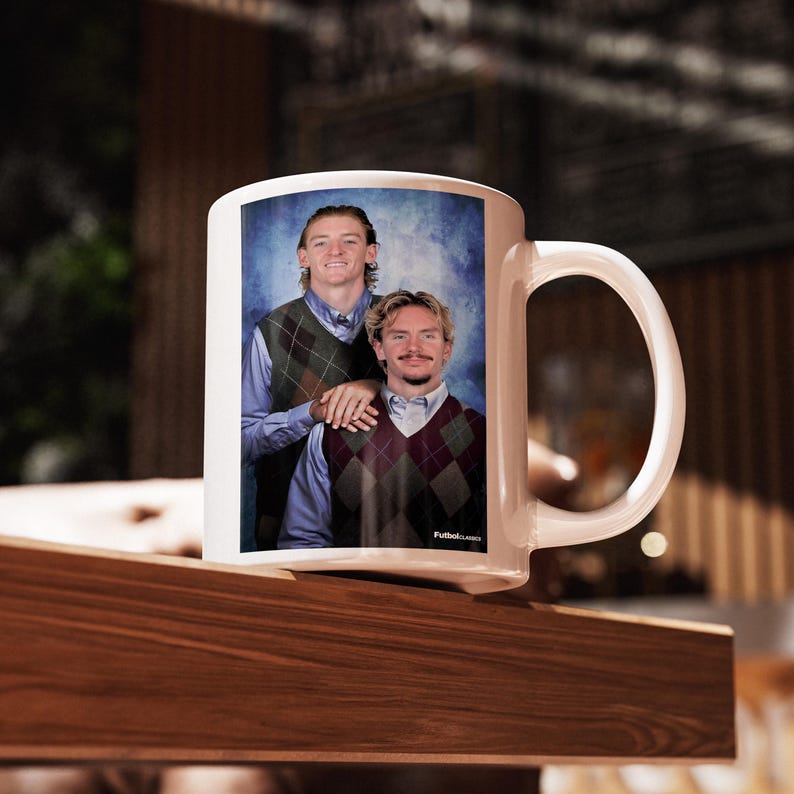 May include: White ceramic mug featuring a portrait of two people wearing argyle vests and collared shirts. The mug has a large handle and the text "Futbol Classics" is visible on the bottom right of the image.