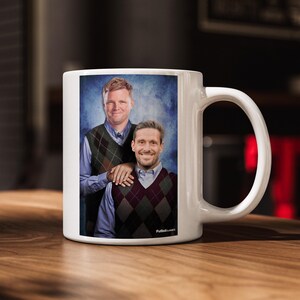 Puede incluir: Taza de cer&aacute;mica blanca con un retrato de dos hombres con chalecos de rombos sobre un fondo azul. La taza tiene un asa curva y el texto "Futbol Classics" est&aacute; impreso en el lateral.