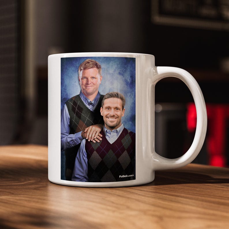 Puede incluir: Taza de cer&aacute;mica blanca con un retrato de dos hombres con chalecos de rombos sobre un fondo azul. La taza tiene un asa curva y el texto "Futbol Classics" est&aacute; impreso en el lateral.