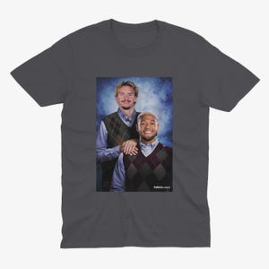 May include: Dark gray t-shirt featuring a portrait of two men in argyle vests and collared shirts. The image has a vintage aesthetic. The text "Futbol Classics" is visible in the bottom right corner.