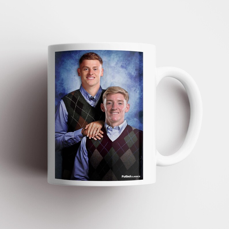 May include: White ceramic mug featuring a portrait of two people in argyle vests and collared shirts against a blue background. The mug has a white handle and the text "FutbolClassics" is visible.