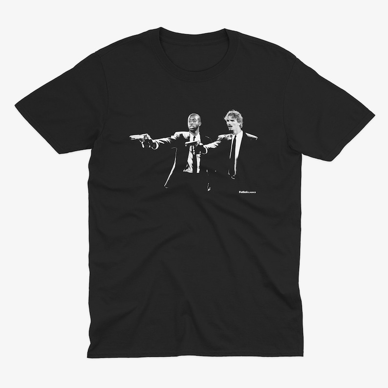 Pu&ograve; includere: T-shirt nero con grafica in bianco e nero di due uomini in giacca e cravatta che puntano pistole. L'immagine &egrave; un'illustrazione stilizzata, con le figure in una posa dinamica. La maglietta &egrave; in stile girocollo classico.
