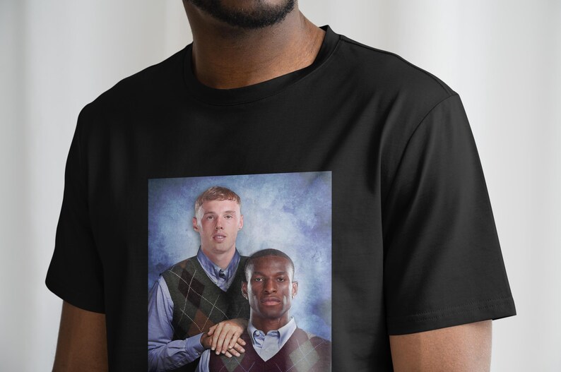 May include: A black t-shirt with a printed image of two men in a school photo. The men are wearing sweaters with a diamond pattern. The background is a blue and white gradient.