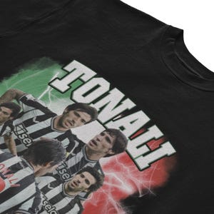 Puede incluir: Camiseta negra con un estampado gr&aacute;fico de jugadores de f&uacute;tbol que llevan camisetas a rayas blancas y negras. El texto "TONALI" est&aacute; impreso en blanco sobre un fondo verde, rojo y blanco.