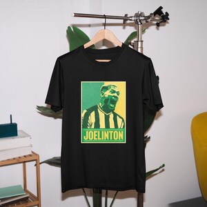 May include: Black t-shirt with a retro-style graphic of a soccer player with the text "JOELINGTON" in yellow.