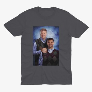 May include: Dark gray t-shirt featuring a framed image of two men. The men are wearing argyle vests over button-down shirts. One man has his hand on the other's shoulder. The background is a blue and white gradient.