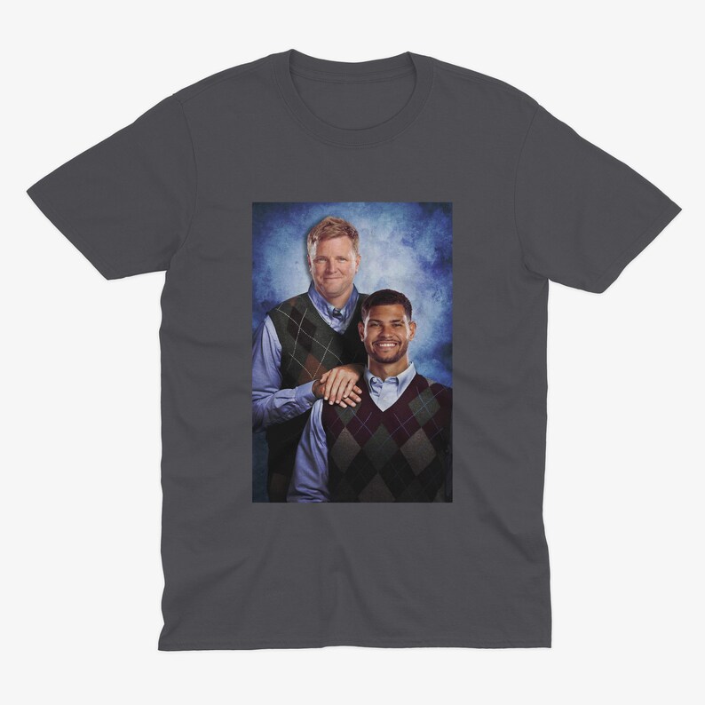 May include: Dark gray t-shirt featuring a framed image of two men. The men are wearing argyle vests over button-down shirts. One man has his hand on the other's shoulder. The background is a blue and white gradient.