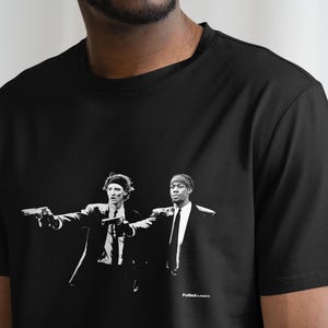 May include: Black t-shirt featuring a black and white graphic of two men in suits holding guns. The image is a stylized illustration. The text "Futbol Classics" is printed at the bottom of the graphic.