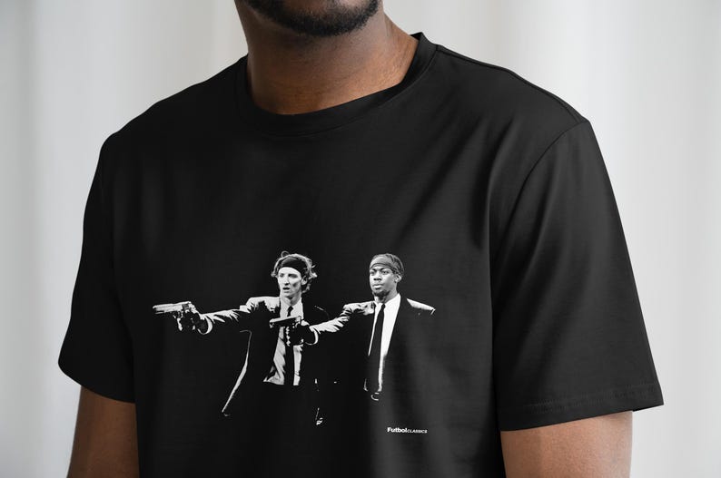 May include: Black t-shirt featuring a black and white graphic of two men in suits holding guns. The image is a stylized illustration. The text "Futbol Classics" is printed at the bottom of the graphic.