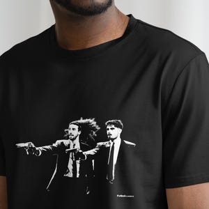 Pu&ograve; includere: T-shirt nero con grafica in bianco e nero di due uomini in giacca e cravatta, ognuno con una pistola. L'immagine ha uno stile noir. Il testo "FutbolLocos" &egrave; in basso.