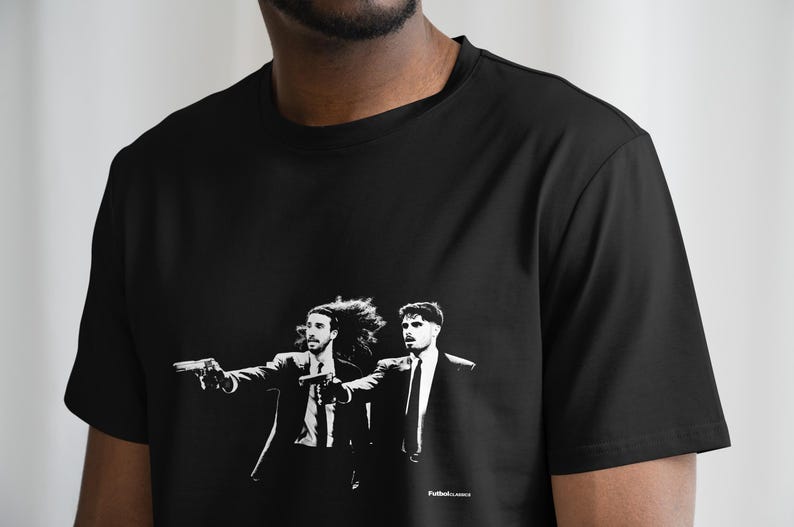 Pu&ograve; includere: T-shirt nero con grafica in bianco e nero di due uomini in giacca e cravatta, ognuno con una pistola. L'immagine ha uno stile noir. Il testo "FutbolLocos" &egrave; in basso.