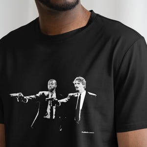 Pu&ograve; includere: T-shirt nero con grafica in bianco e nero di due uomini in giacca e cravatta che imbracciano pistole. L'immagine &egrave; un'illustrazione stilizzata. Il testo "Futbol Classics" &egrave; stampato in basso.