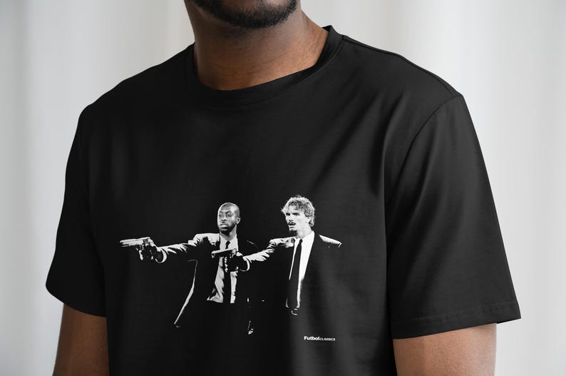 Pu&ograve; includere: T-shirt nero con grafica in bianco e nero di due uomini in giacca e cravatta che imbracciano pistole. L'immagine &egrave; un'illustrazione stilizzata. Il testo "Futbol Classics" &egrave; stampato in basso.